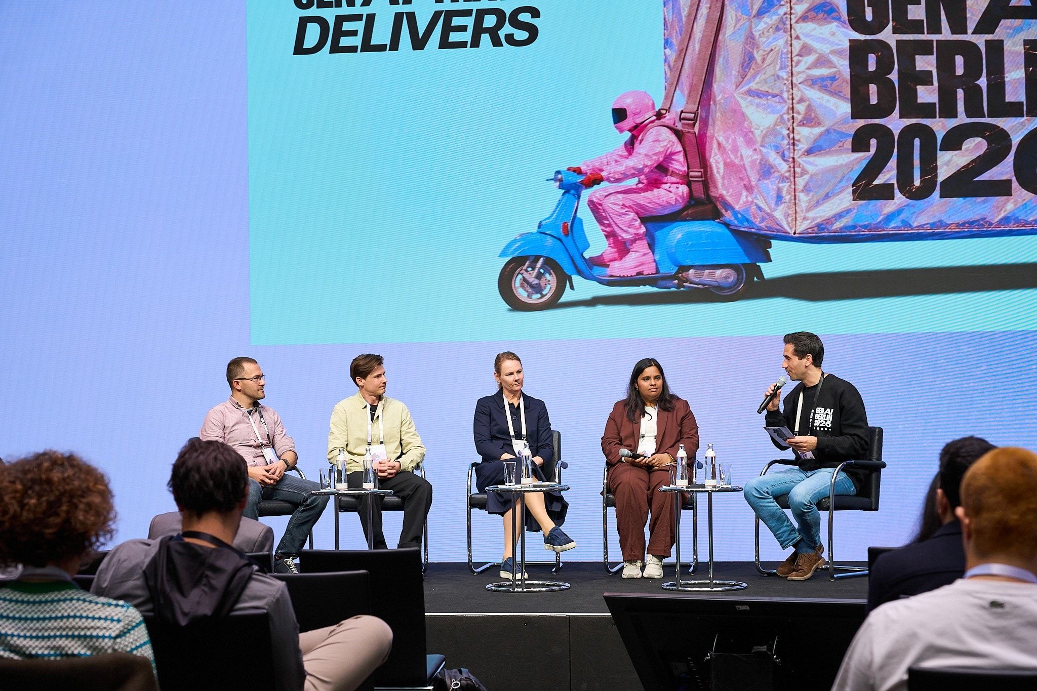 Five panellists seated on stage at GenAI Berlin 2026, with Sasa Fajkovic on the far left and the moderator holding a microphone on the right