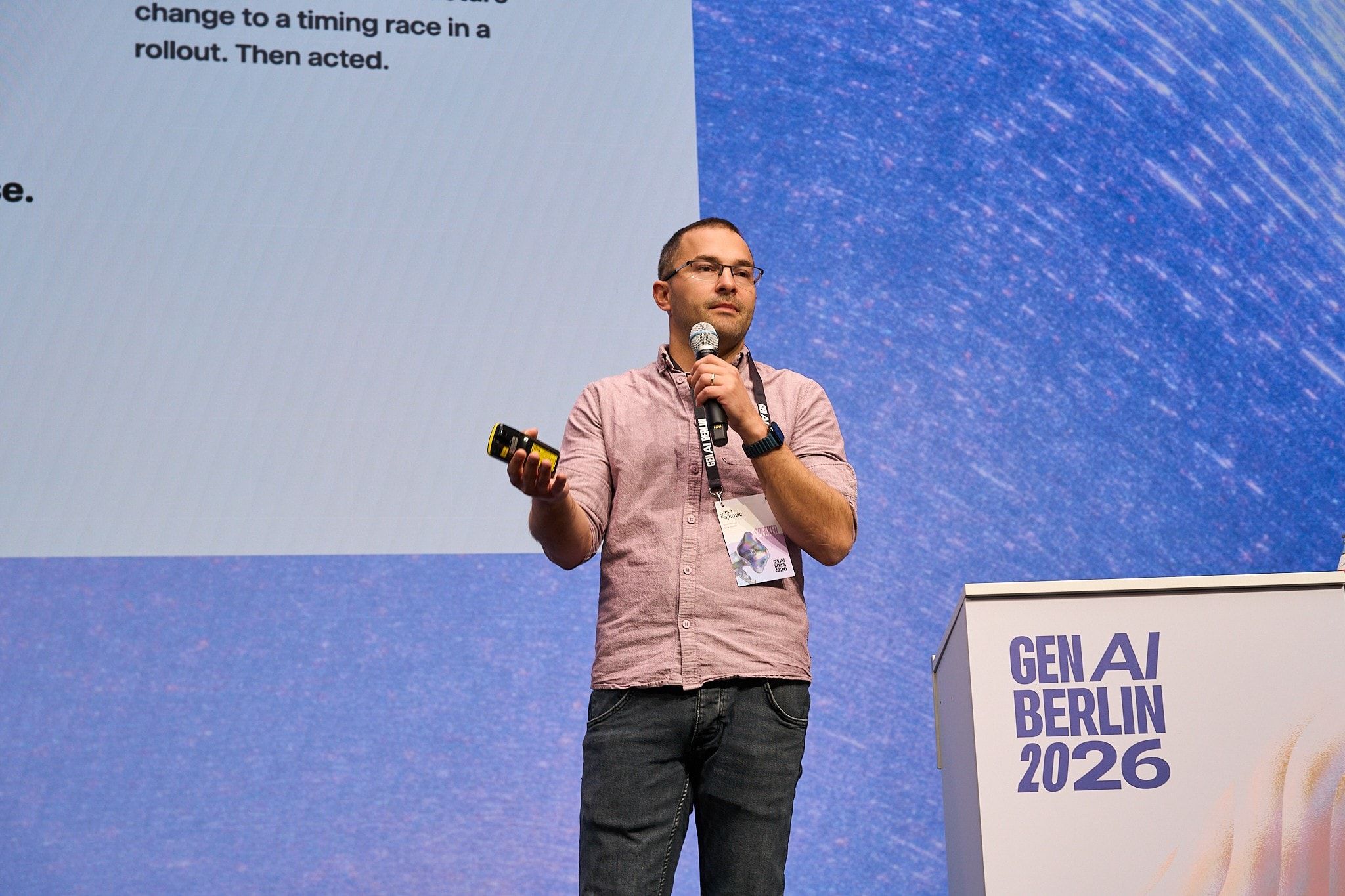 Sasa Fajkovic speaking into a handheld microphone on stage at GenAI Berlin 2026, in front of a large projected slide