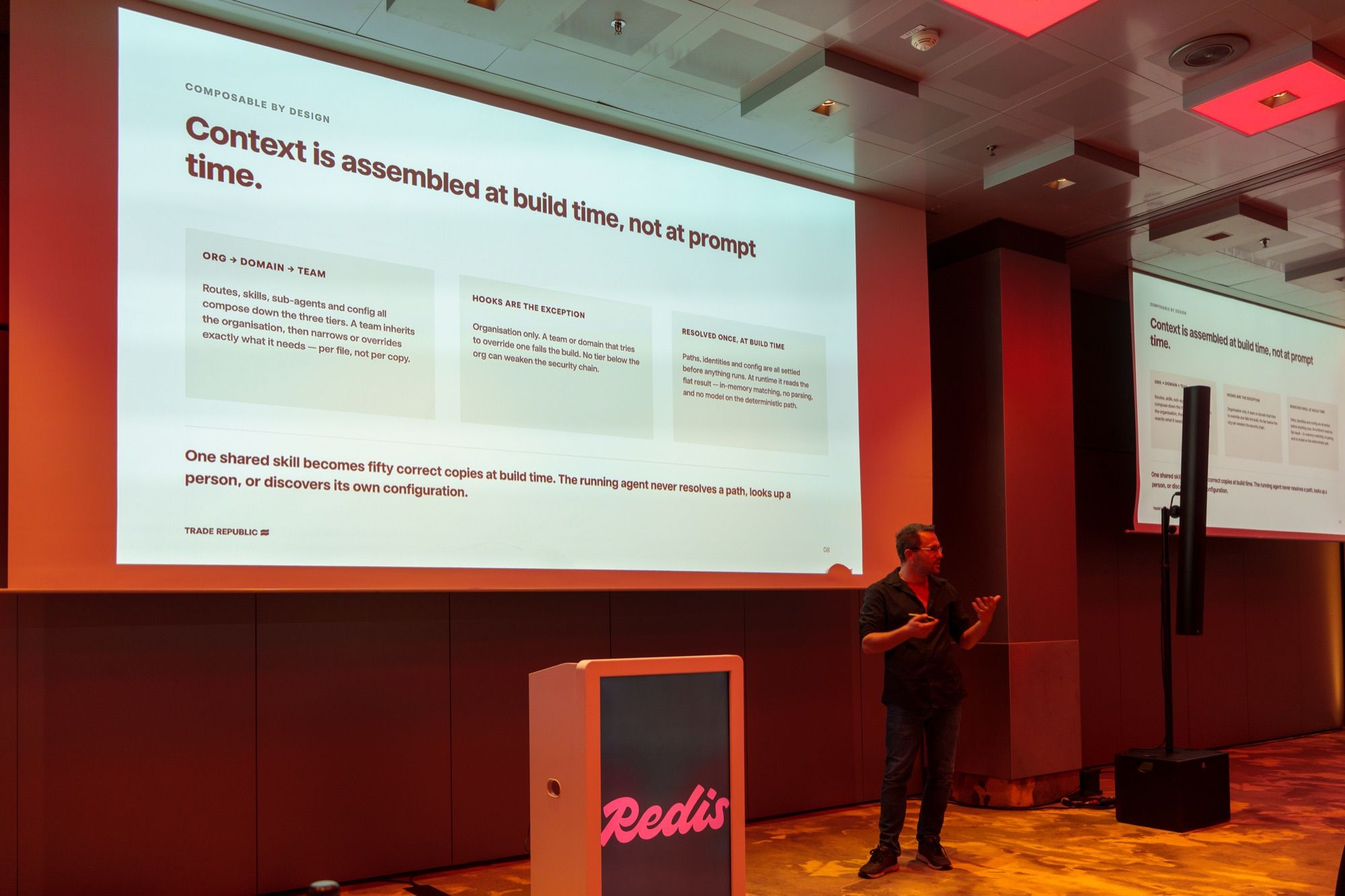 Sasa Fajkovic turning towards the build-time context slide on stage at the Redis AI Roadshow in Munich, with a second screen on the right