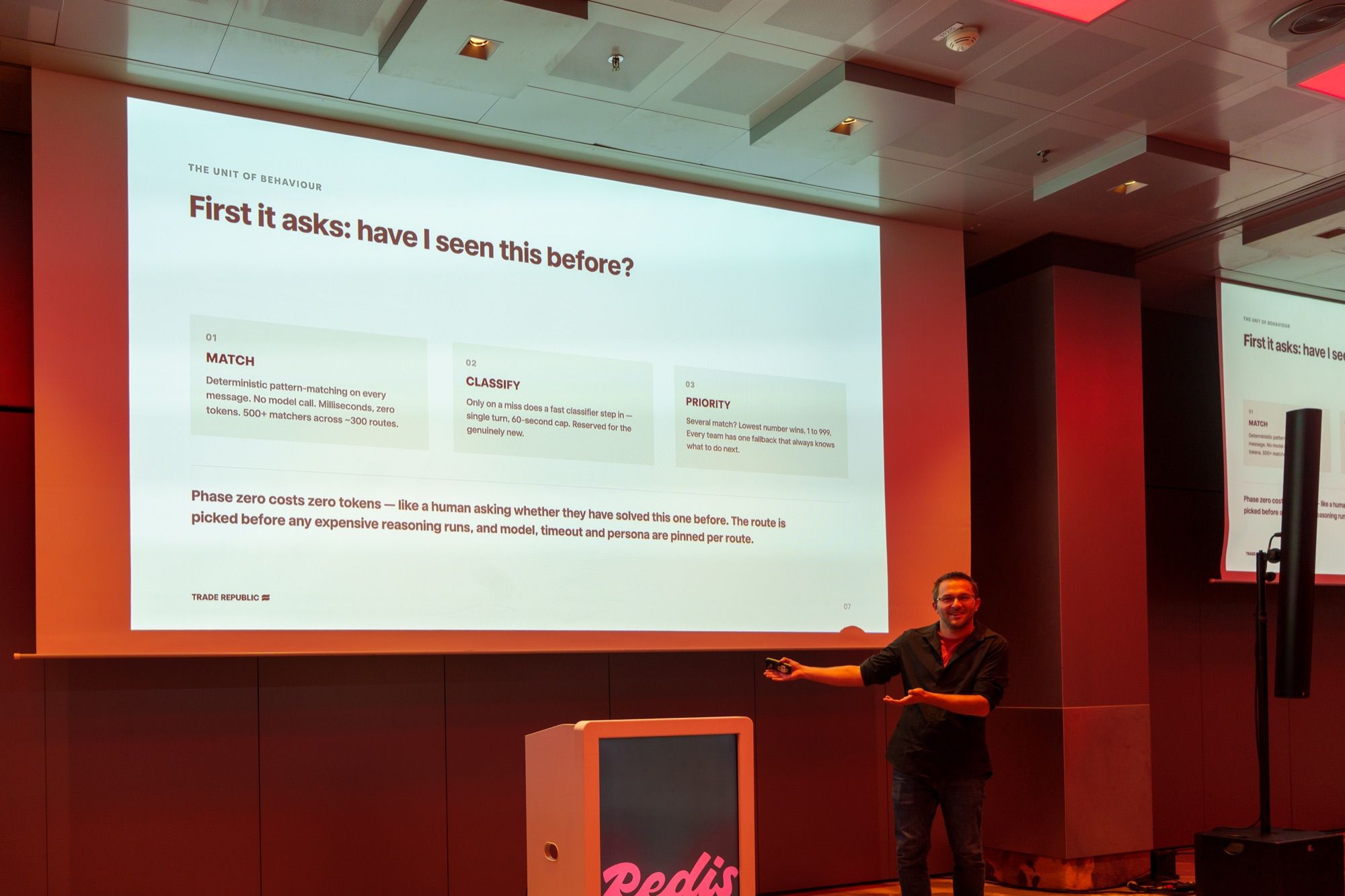 Sasa Fajkovic smiling and gesturing towards a slide titled “First it asks: have I seen this before?” beside the Redis lectern in Munich